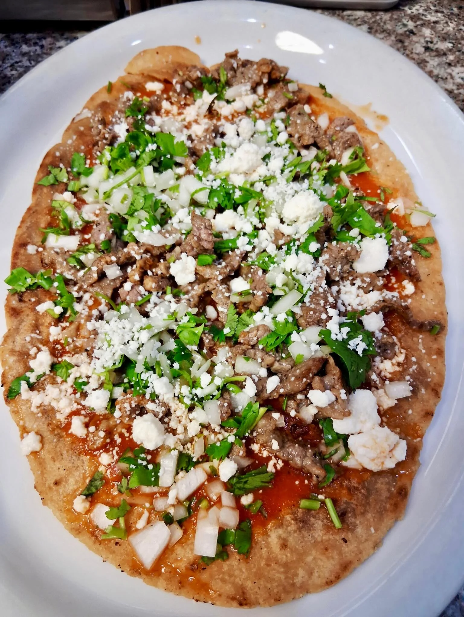 A delicious flatbread topped with seasoned beef, diced onions, fresh cilantro, and crumbled cheese, served on a white plate.