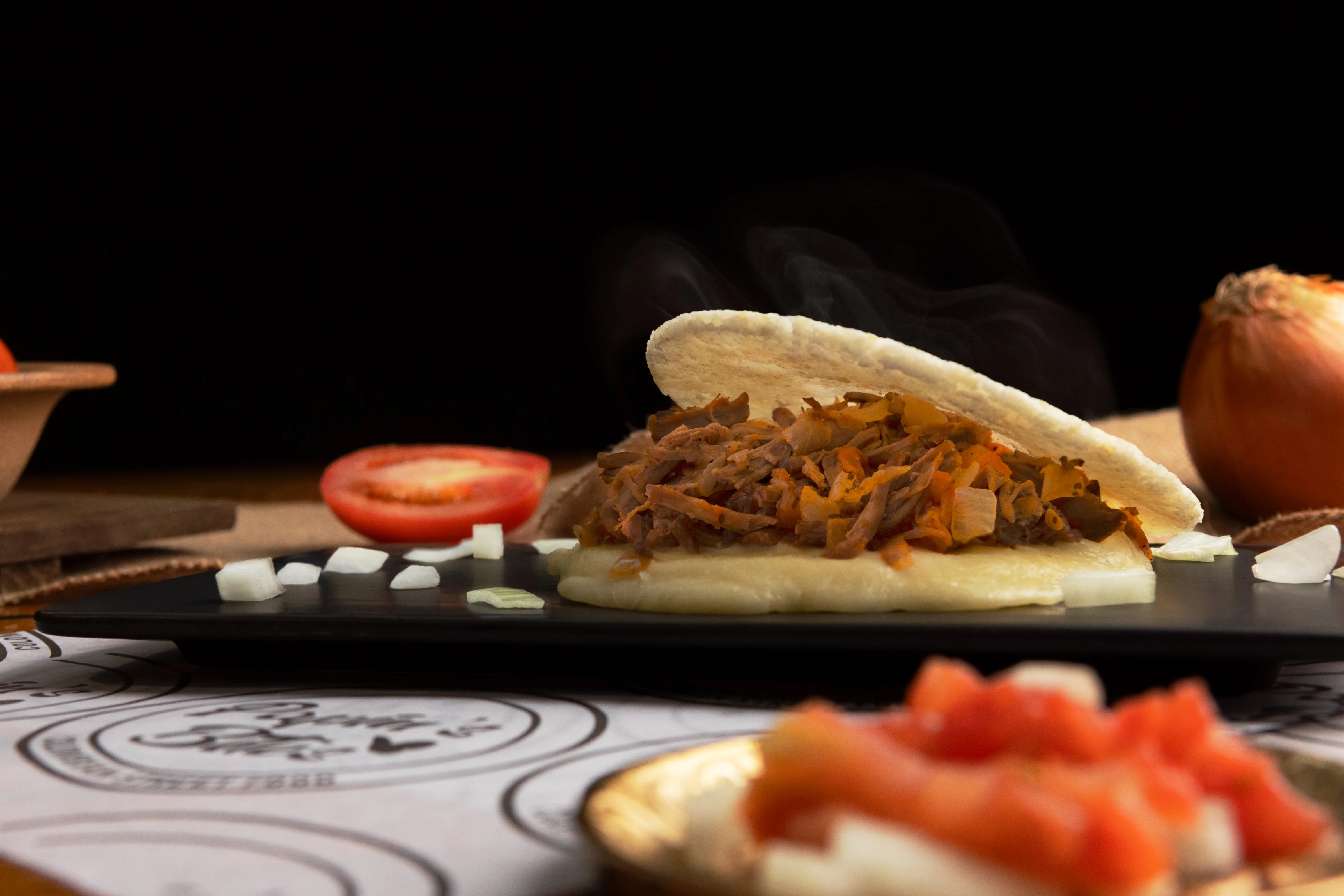 A delicious arepa filled with shredded meat and vegetables, served on a black plate with fresh diced ingredients and a tomato in the background.