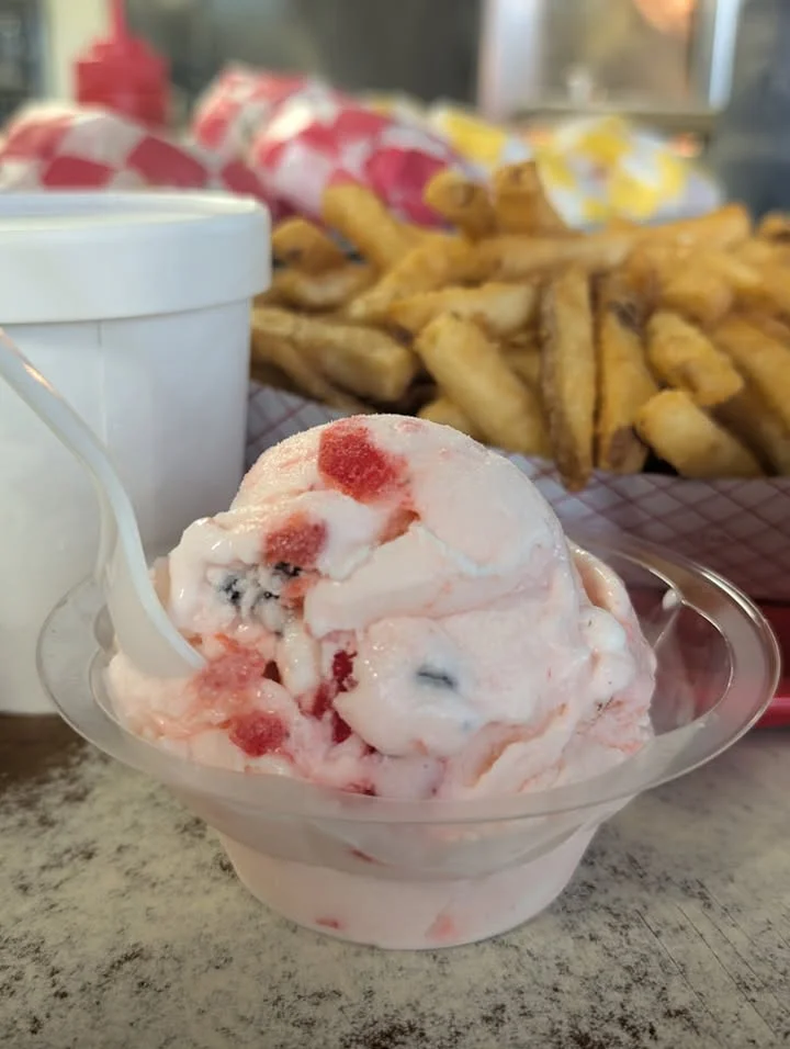 A scoop of strawberry ice cream with bits of fruit, served in a clear cup, alongside fried potatoes in the background.