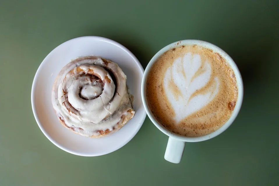 A delicious cinnamon roll topped with icing, served on a white plate alongside a latte with artistic foam design in a contrasting cup.
