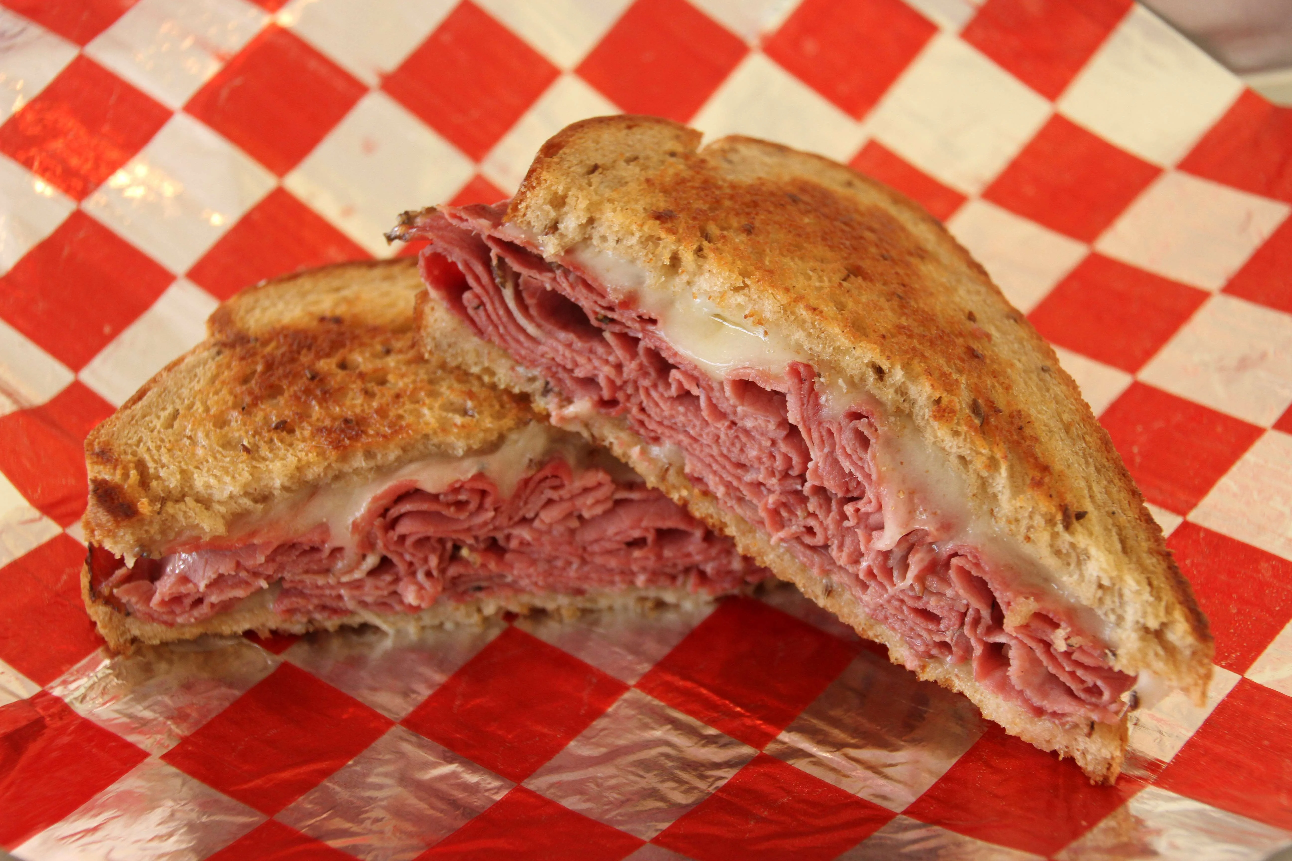Hot pastrami sandwich on toasted rye bread with melted Swiss cheese, served on red and white checkered deli paper.
