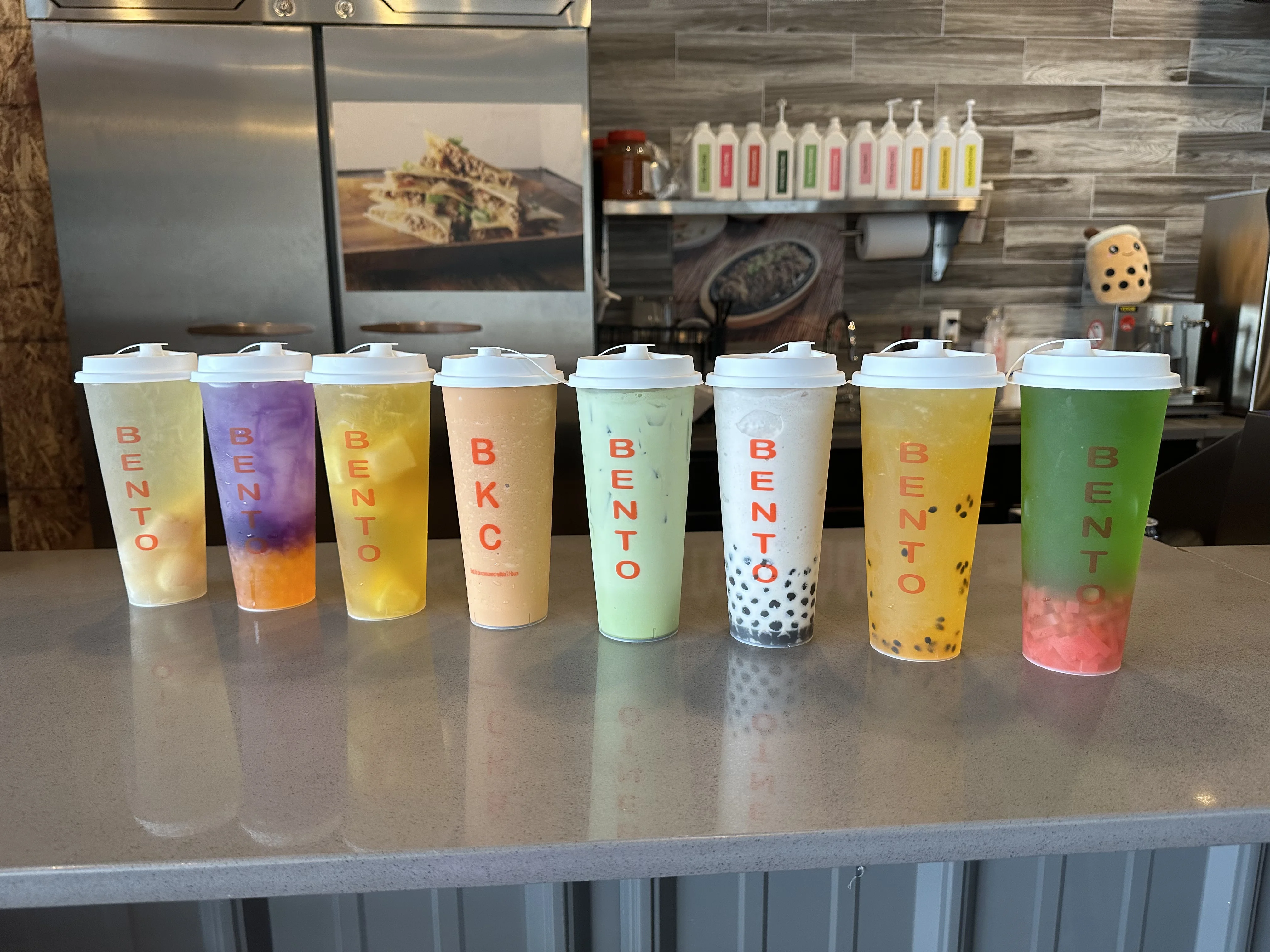 Assortment of colorful tea drinks in tall cups labeled 'BENTO' at Bento Kitchen Cravings, featuring various flavors with fruit pieces, tapioca pearls, and vibrant layers, displayed on a counter in a modern restaurant setting.