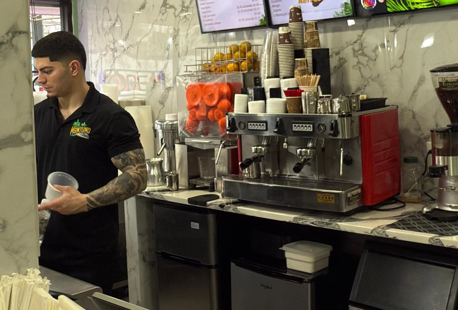 The image displays the service counter of a café with a marble-patterned countertop. On it, there’s a "Tips" jar, straws, and a small payment terminal. Behind are digital menu screens listing drink and food options, alongside a coffee machine and grinder.
