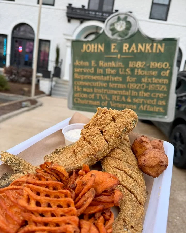 A platter of fried fish and sweet potato fries, with a side of dipping sauce, placed in front of a historical marker.