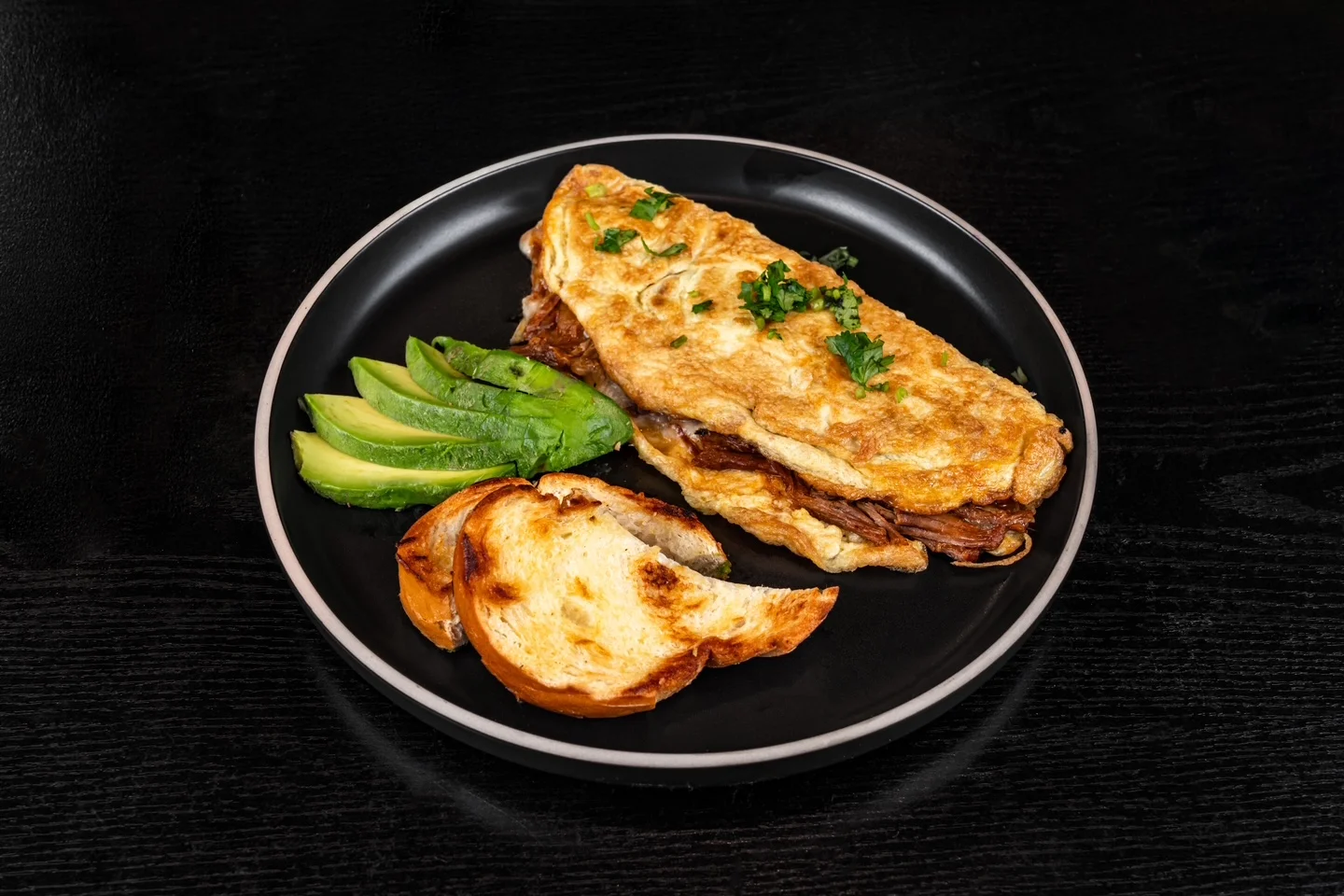 A plate featuring a hearty omelette filled with meat, accompanied by avocado slices and toasted bread.