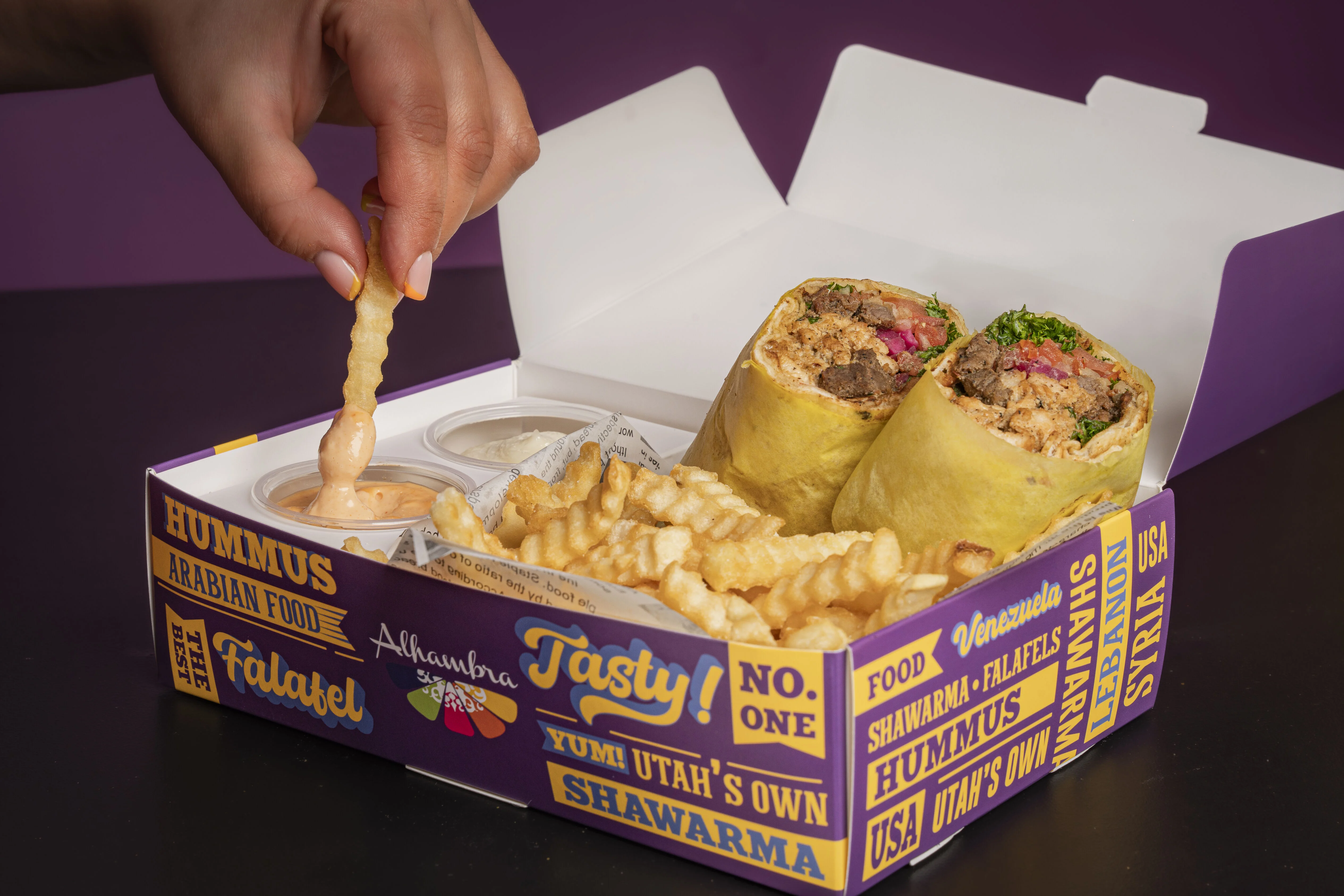 A hand dipping a fry into sauce next to wraps and crinkle fries in a takeout box.