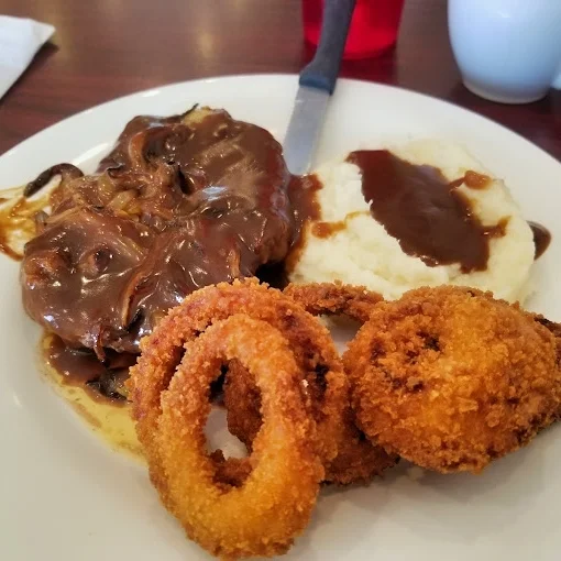 A plate featuring a tender meat cut topped with rich onion gravy, served with a side of mashed potatoes and crispy onion rings.