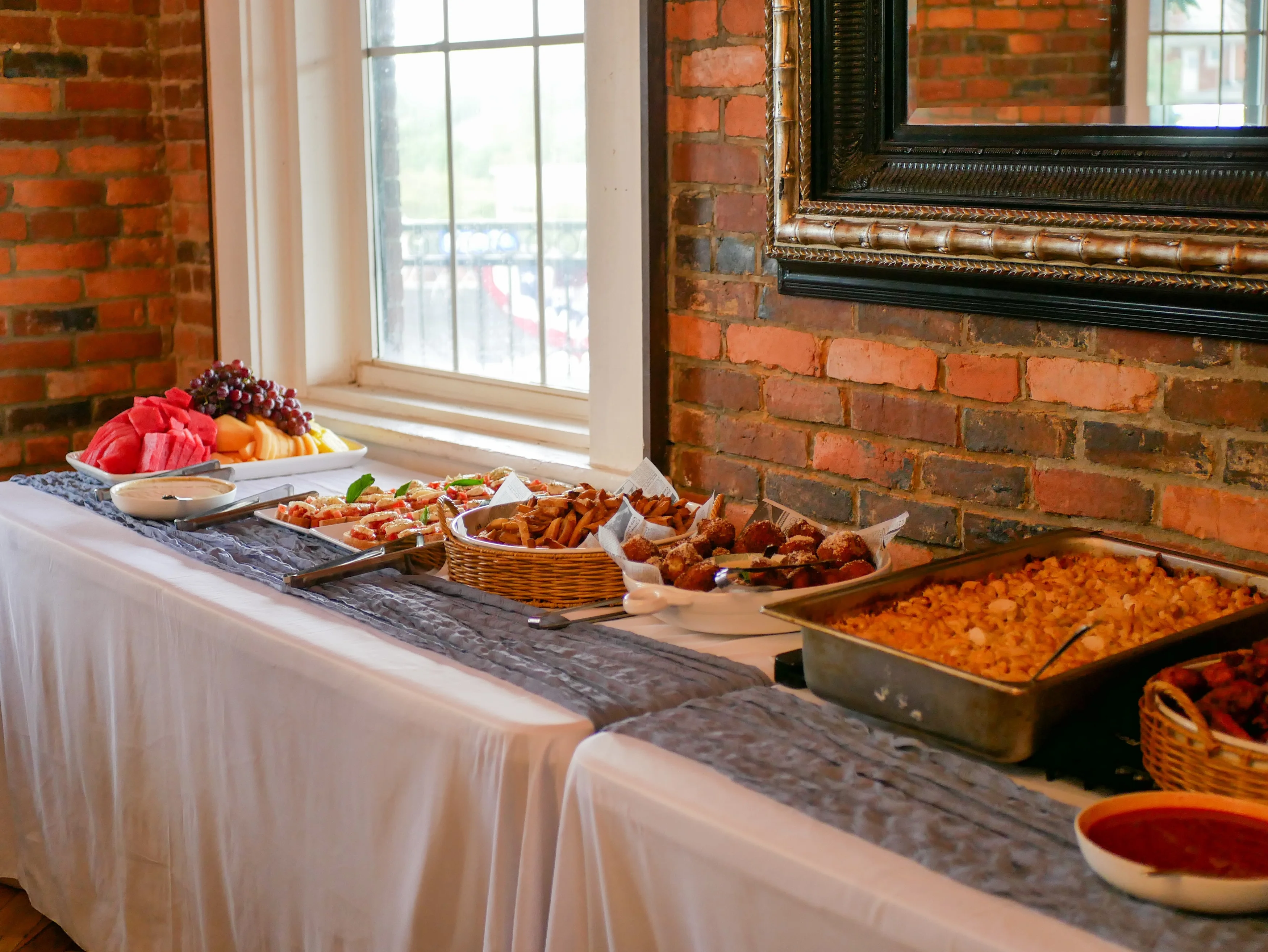 A buffet table featuring a variety of fruits, appetizers, and dishes including watermelon, grapes, and snacks. The table is set against a rustic brick wall.