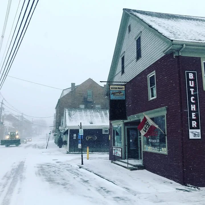 A snowy street scene featuring a red butcher shop on the right and a seafood market across the street, with snow accumulating on the ground.