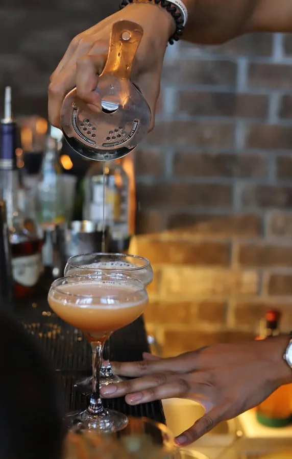 A bartender pours a cocktail from a strainer into two elegant coupe glasses, set against a backdrop of various bottles.