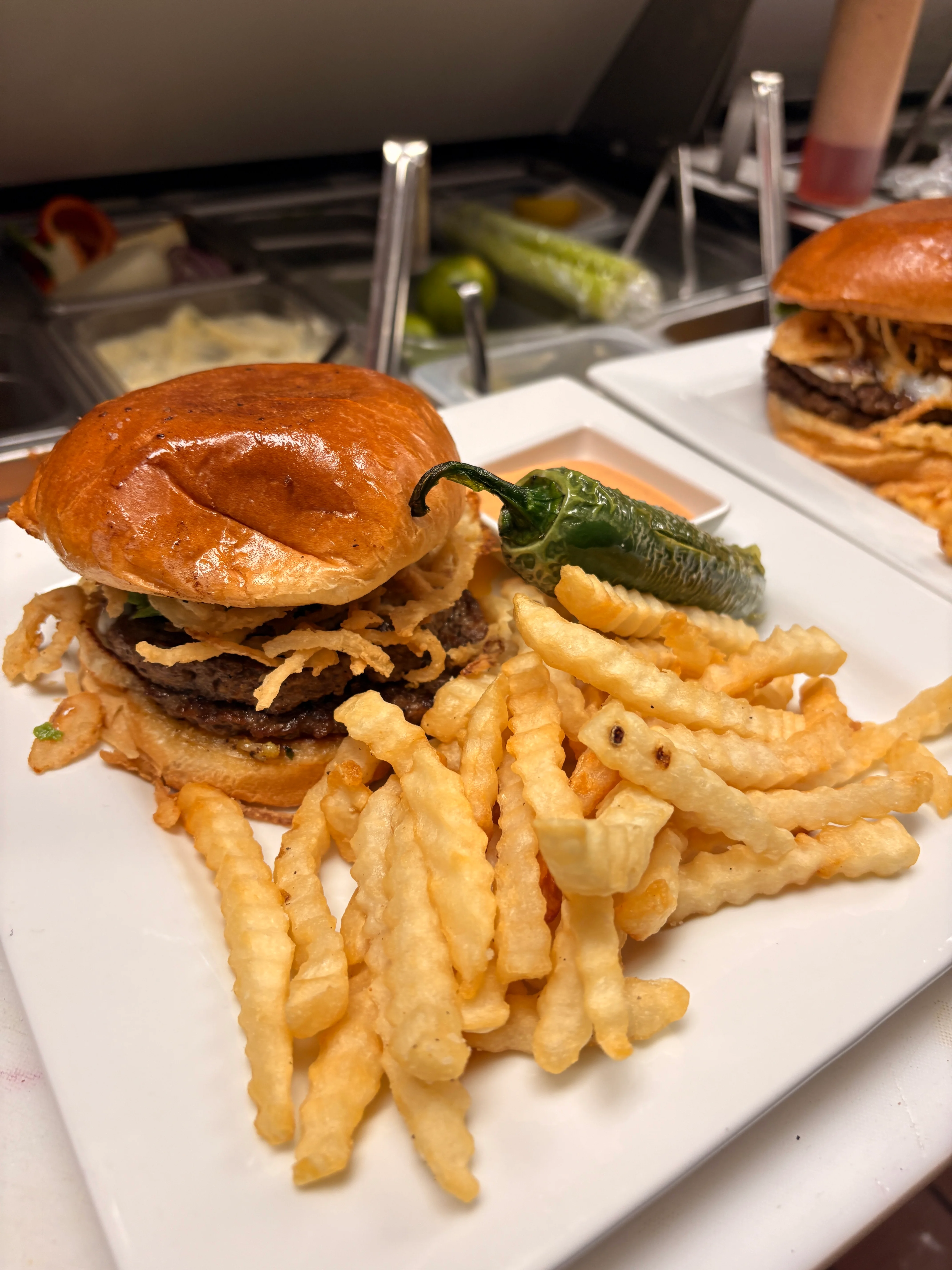 A gourmet burger with crispy onion strings, served alongside golden crinkle fries and a jalapeño pepper.