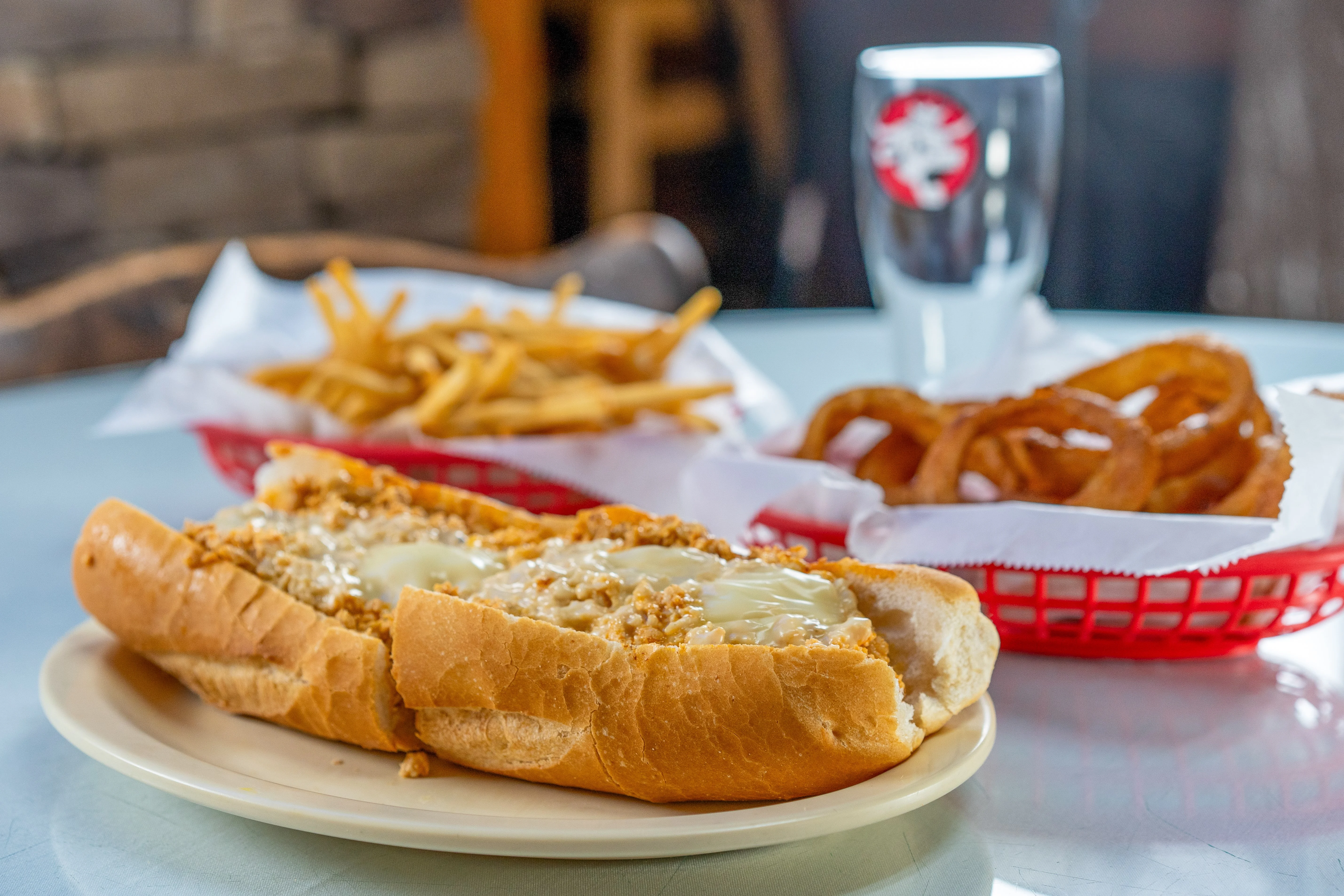 Buffalo chicken cheesesteak topped with melted cheese in a toasted roll, served with golden fries and crispy onion rings in red baskets