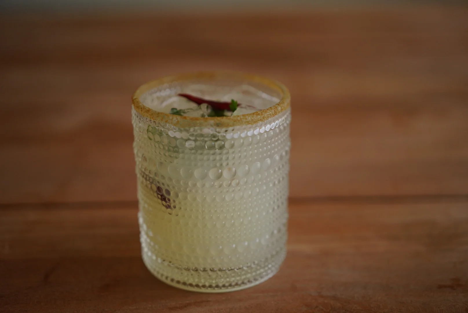 A textured glass cup filled with a light-colored beverage, garnished with a chili and herbs. The cup is placed on a wooden surface.