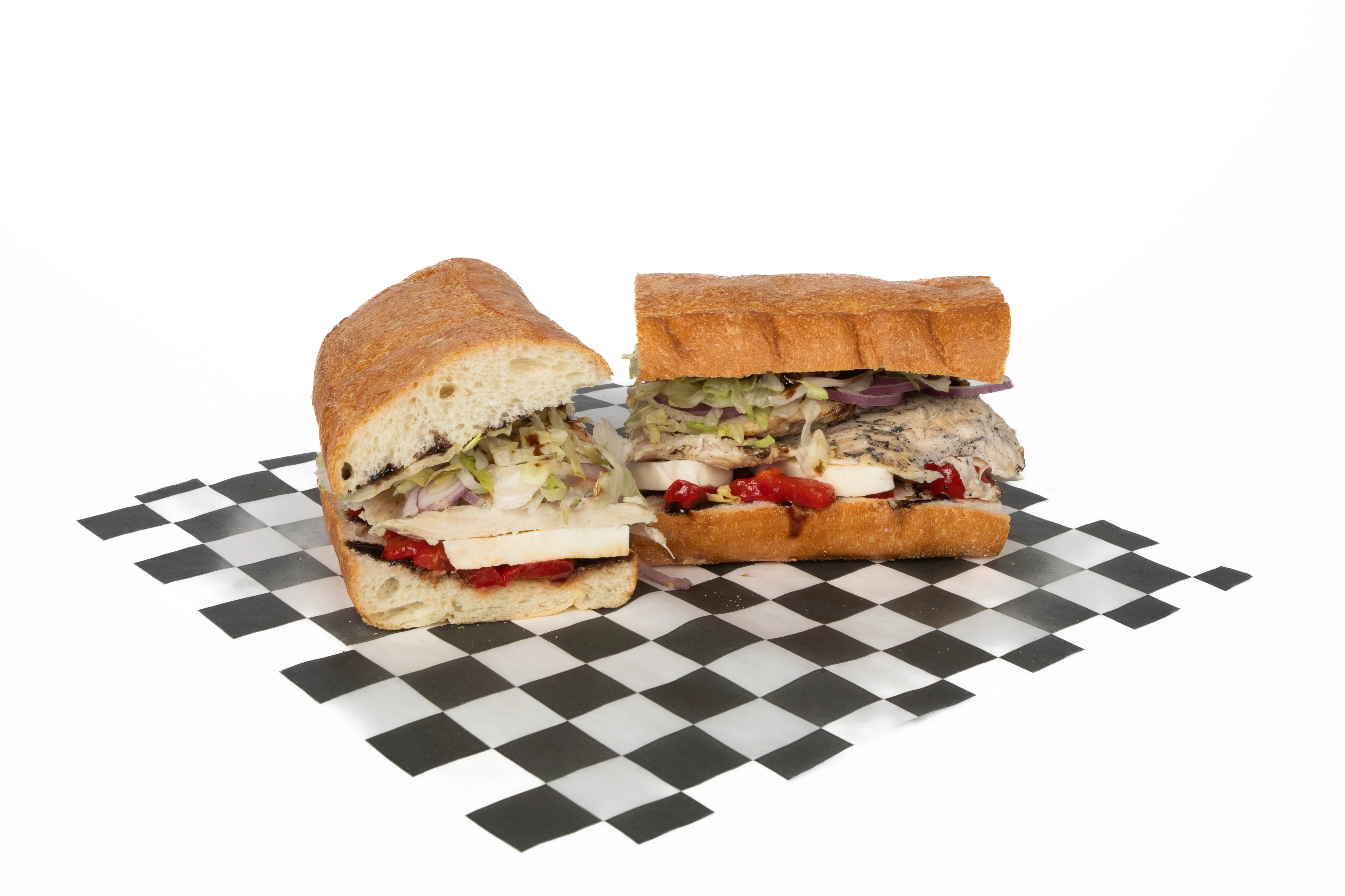 Two halves of a grilled sandwich filled with roasted chicken, vegetables, and condiments, placed on a checkered surface.