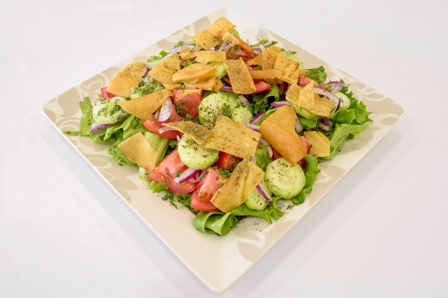 A fresh salad featuring a mix of greens, diced tomatoes, cucumber slices, and red onions, topped with crispy tortilla strips and sprinkled with herbs. Served on a decorative plate.