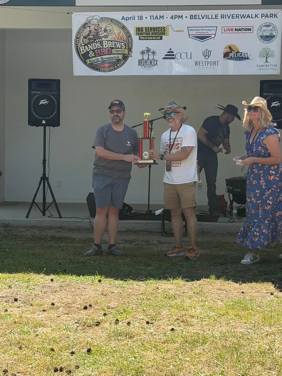 A group of people standing in front of a food trailer named 'Smokin with Bigg'. Some individuals are holding trophies, and the setting has trees in the background.