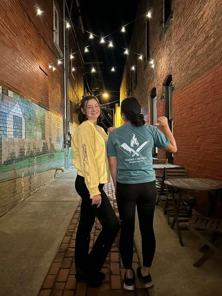 Two individuals posing in a dimly lit alleyway, adorned with string lights. One is wearing a yellow sweatshirt while the other sports a teal shirt.