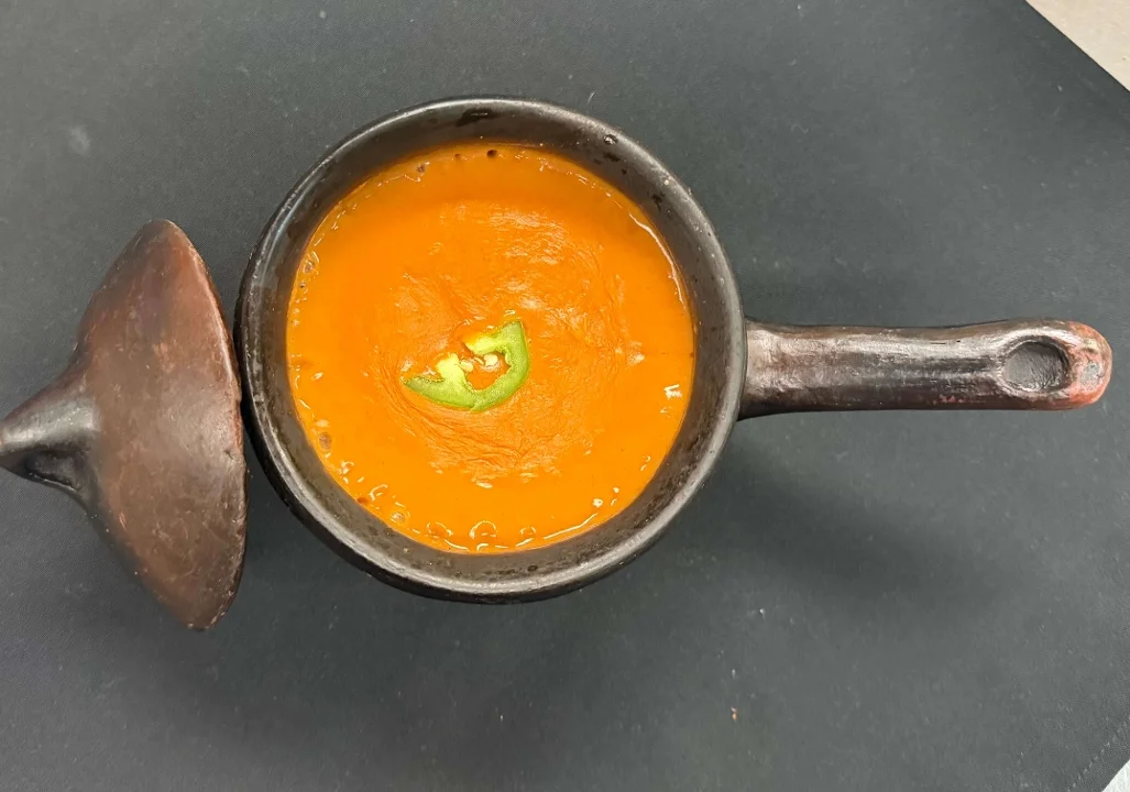 A spicy orange soup in a traditional clay pot, garnished with a green slice.