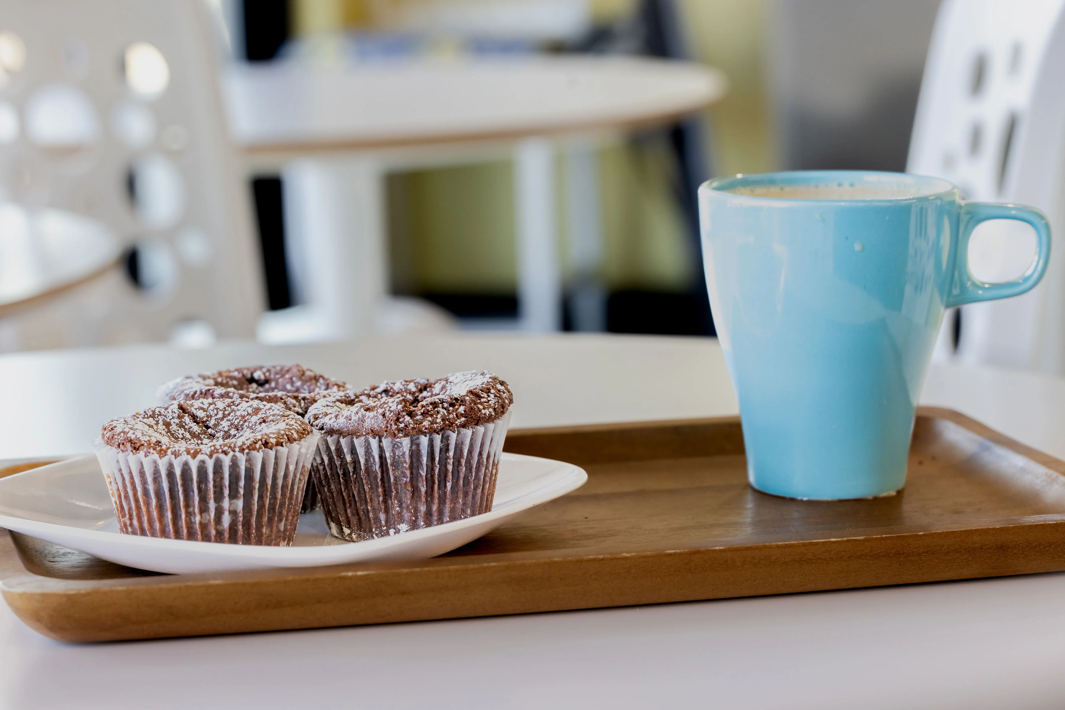 A wooden tray holds three chocolate muffins dusted with powdered sugar alongside a steaming blue cup of coffee.