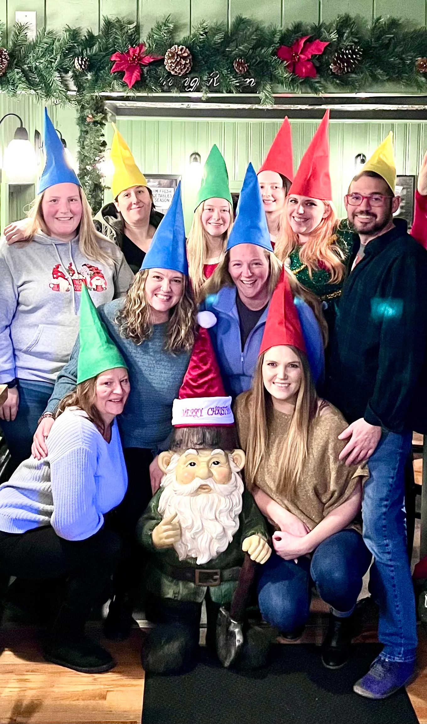 A group of people wearing colorful cone-shaped hats and posing with a cheerful garden gnome statue. The setting includes festive decorations in the background.