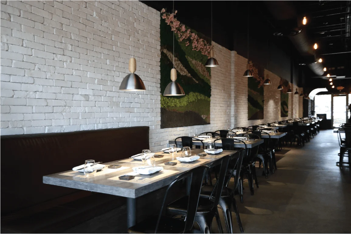 A restaurant dining area featuring a row of black tables and chairs along a white brick wall, decorated with moss wall art and industrial pendant lighting.