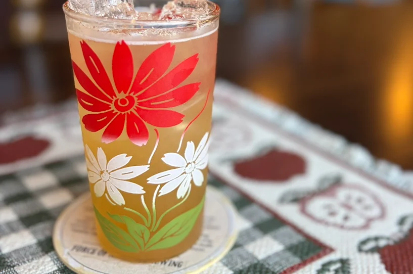 Two glasses of craft beer on coasters. The left glass is dark with a foamy top, labeled 'Antares' and 'Belgian Quadrupel'. The right glass is bright red, labeled 'Bête Rouge' and 'French Farmhouse Saison w/ Beets'.