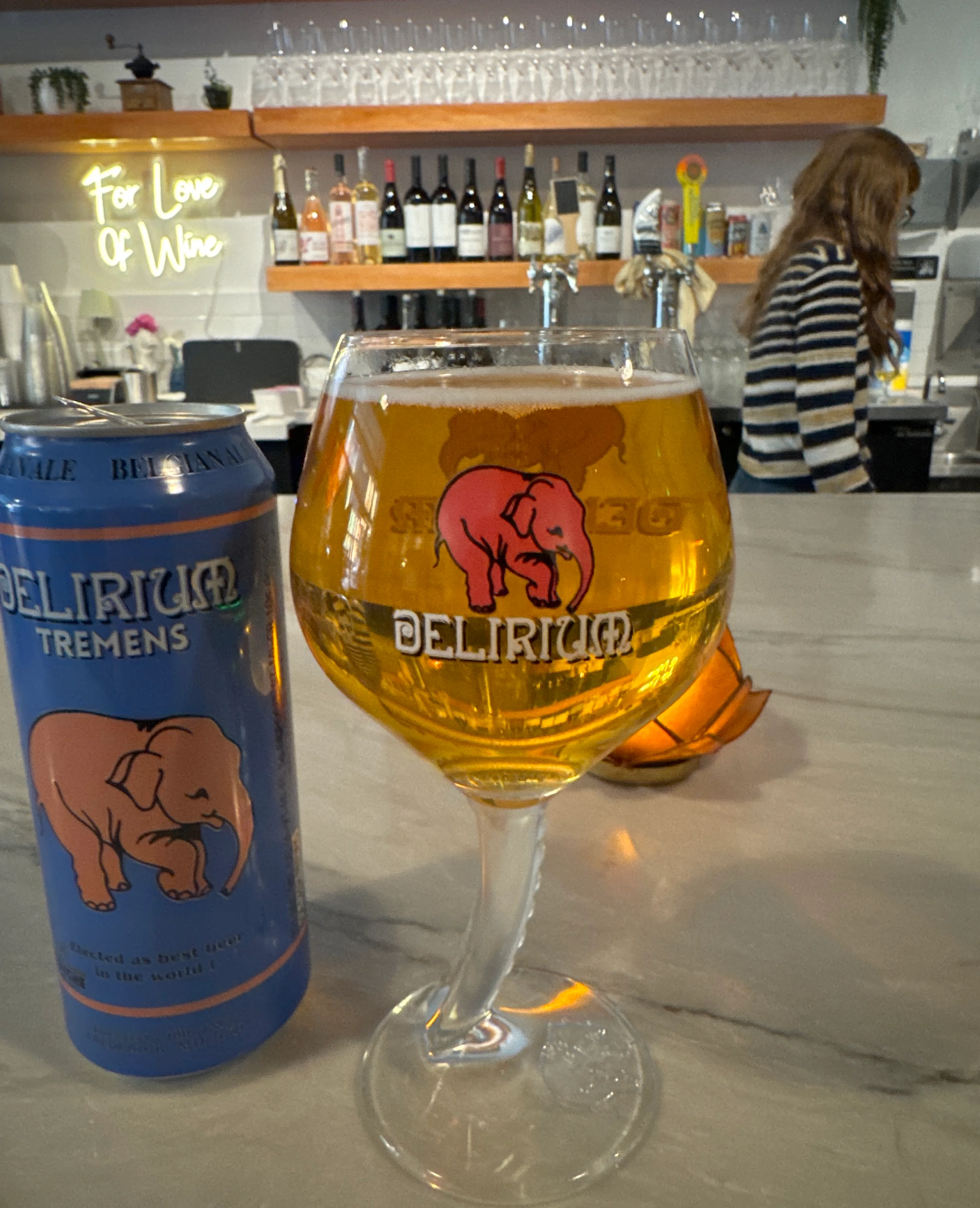 A glass of Delirium Tremens beer next to its can on a bar counter.