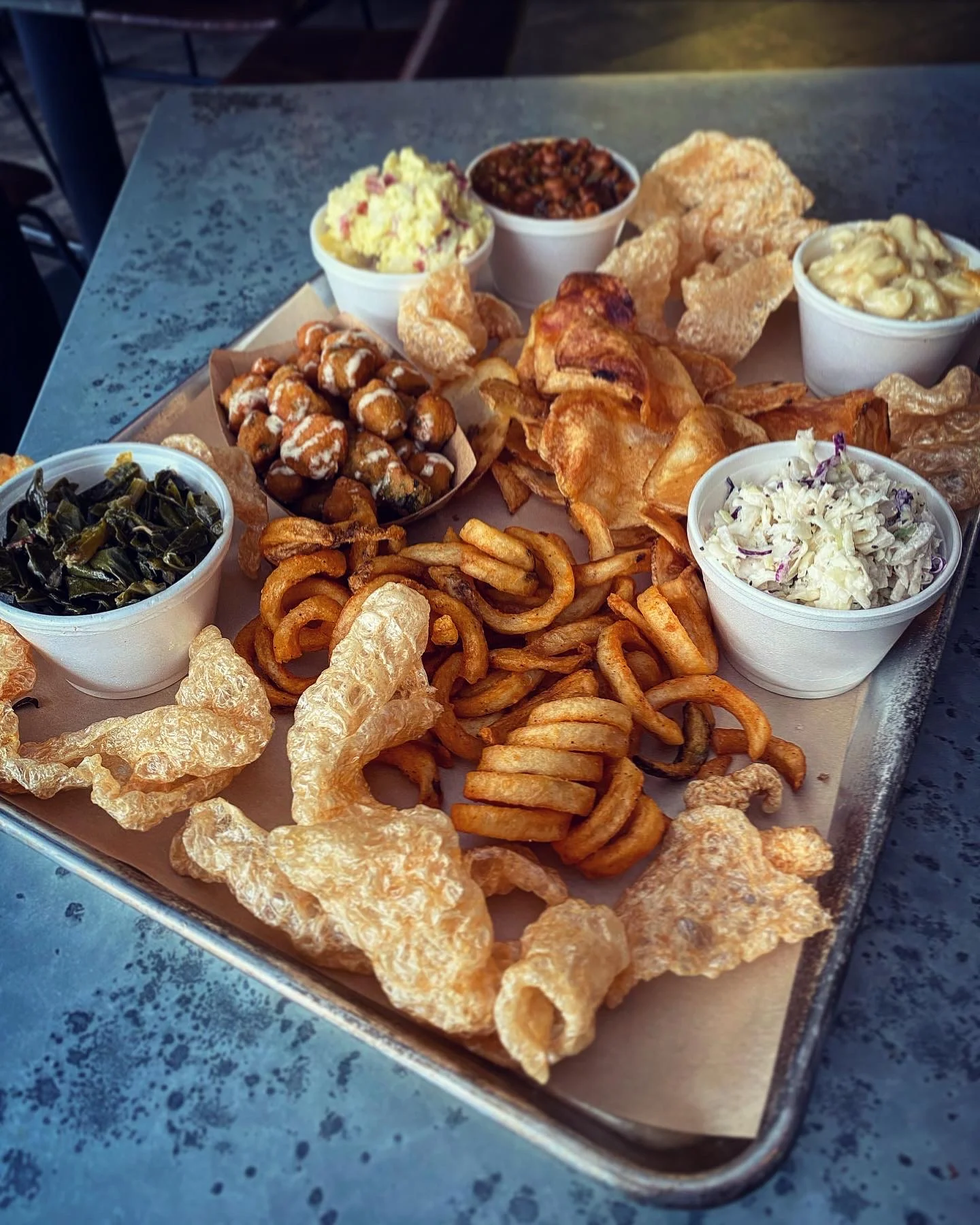 A large tray filled with various fried foods including pork rinds, curly fries, and several small cups of different sides such as coleslaw and macaroni.