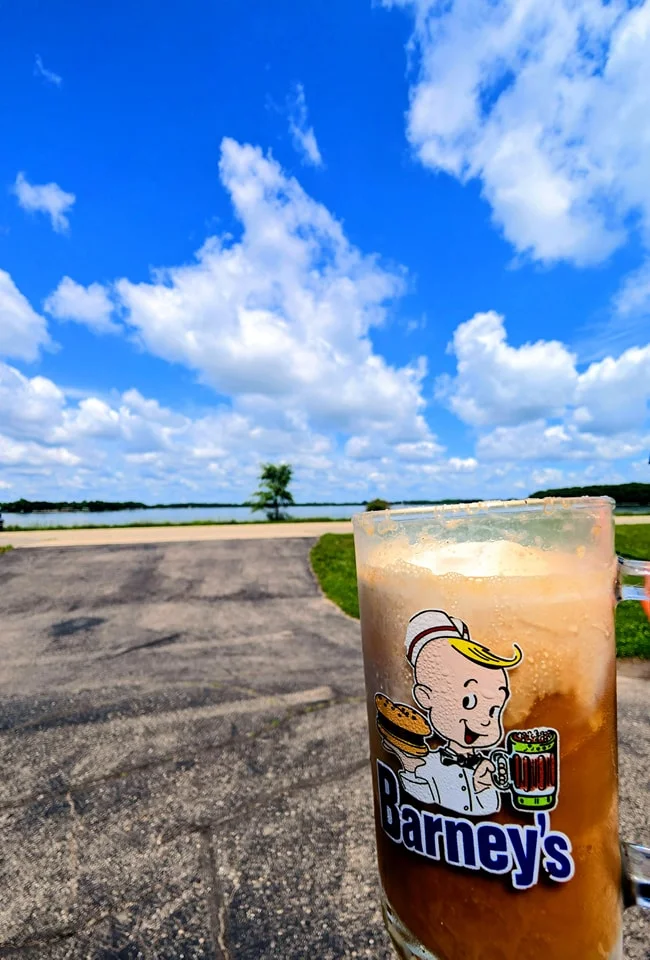 A frosty glass of root beer sits in the foreground with a scenic view of a lake and a blue sky filled with fluffy clouds in the background.