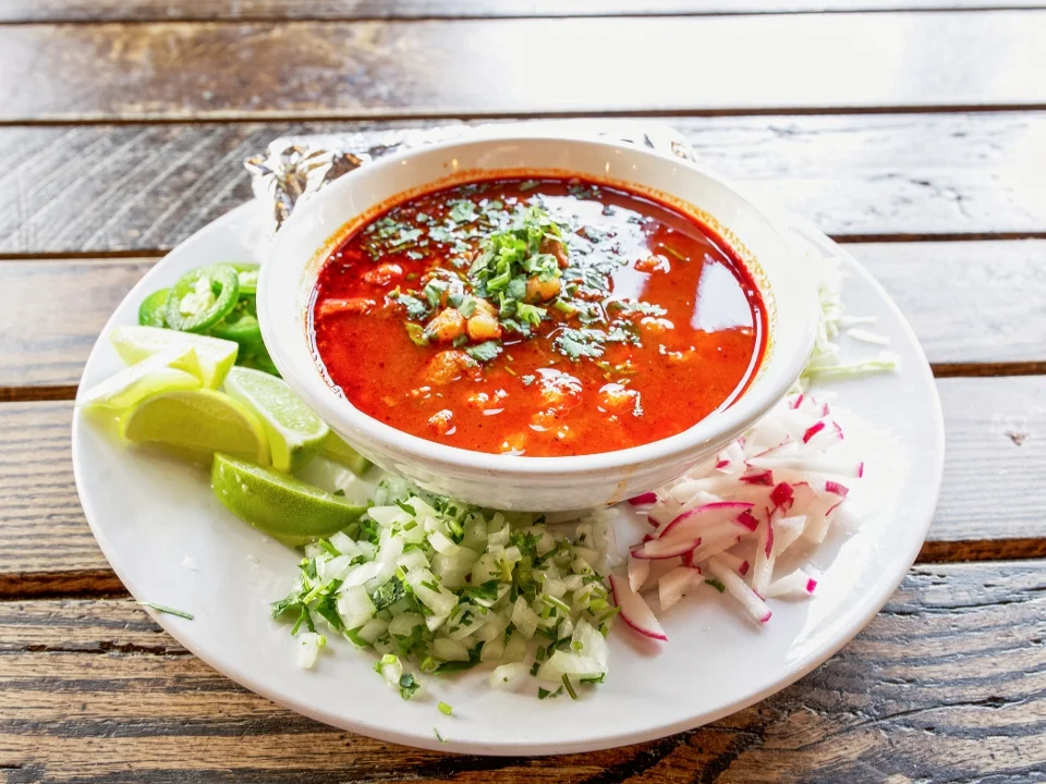 A bowl of red soup garnished with diced ingredients, served on a plate with lime wedges, chopped onions, and radishes.