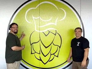 Two chefs posing by a large hop cone logo in a brewery