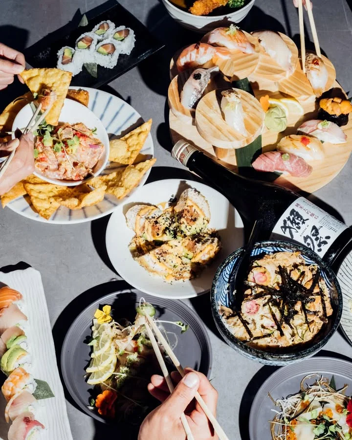 A variety of sushi and Japanese dishes displayed on a table, including sushi rolls, sashimi, fried dumplings, and a bowl of rice. Hands are seen using chopsticks.