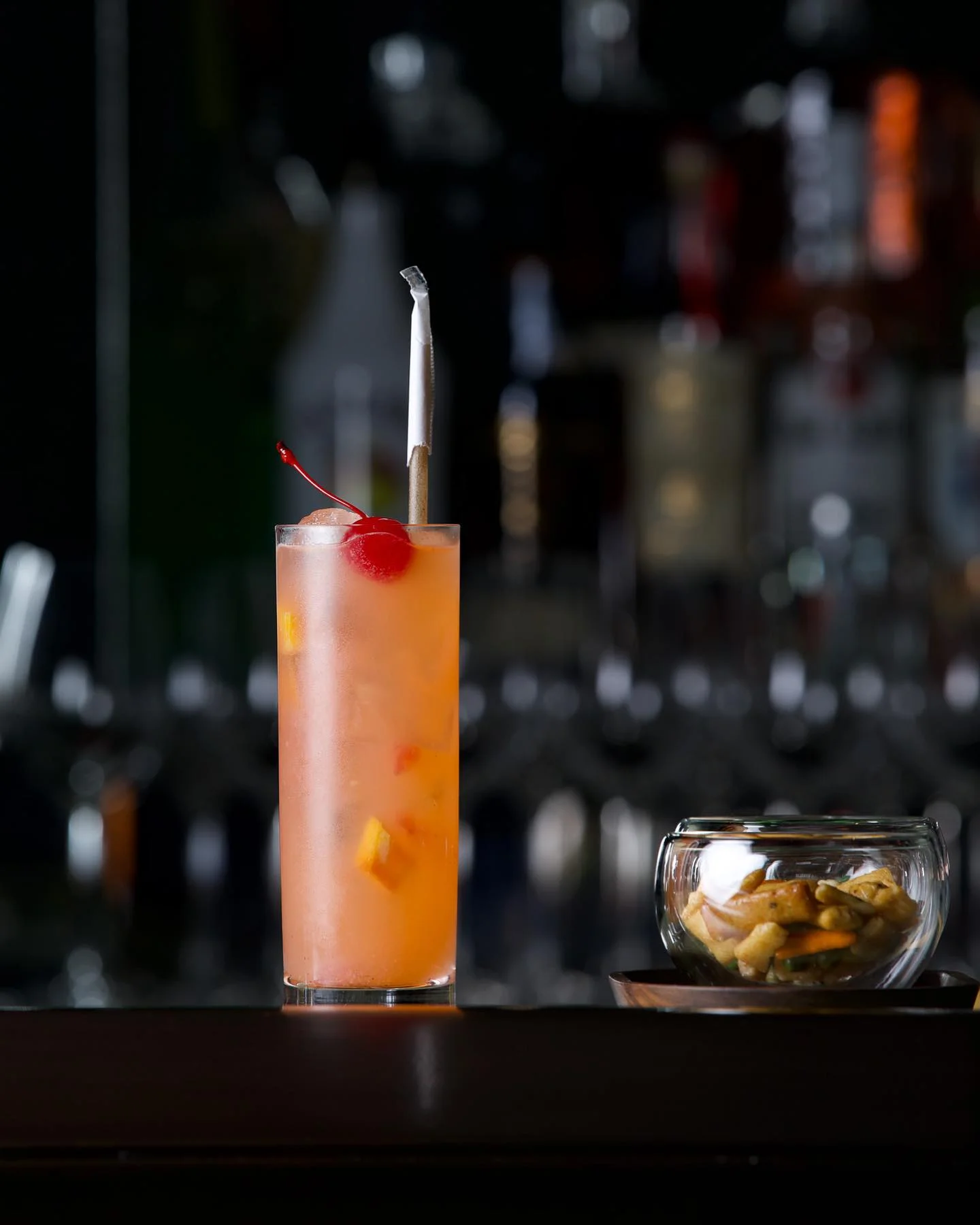 A tall, chilled orange-pink cocktail served in a highball glass containing ice chunks and citrus slices, topped with a bright red maraschino cherry and a paper-wrapped straw, positioned next to a small glass bowl filled with bar snacks against a dark, blurred bar background.