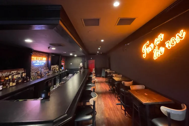 Warmly lit bar interior of The Fox's Den at The Fox Restaurant & Brewery Carson City, featuring glowing 'Fox on the Rocks' neon sign, brick walls, and cozy seating