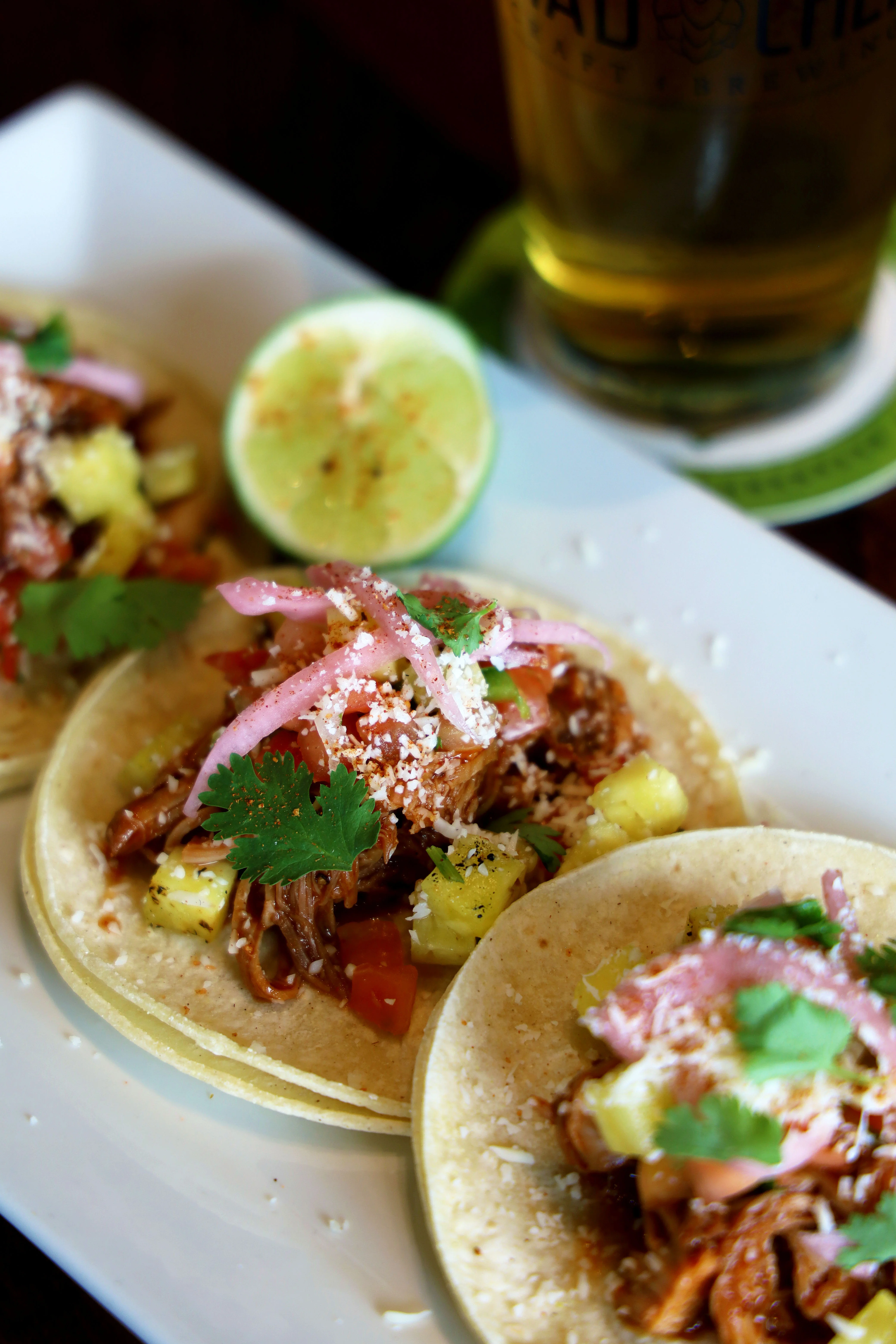 Three tacos with pulled meat, pineapple, and lime on a plate.