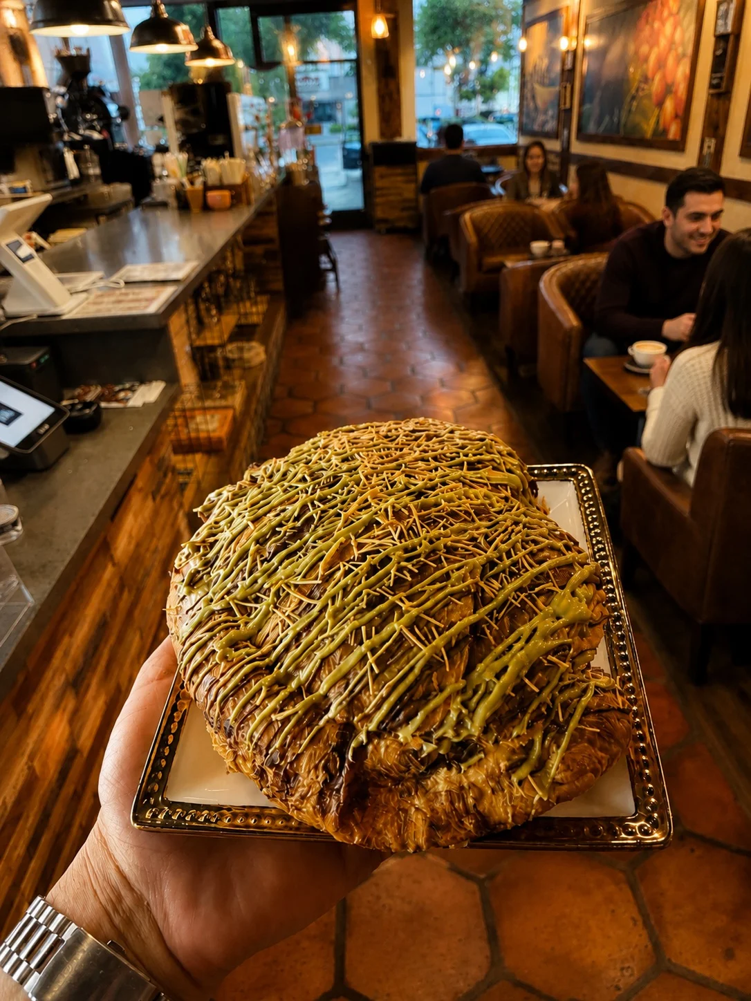 A hand holding a large pastry drizzled with green and brown icing in a café.