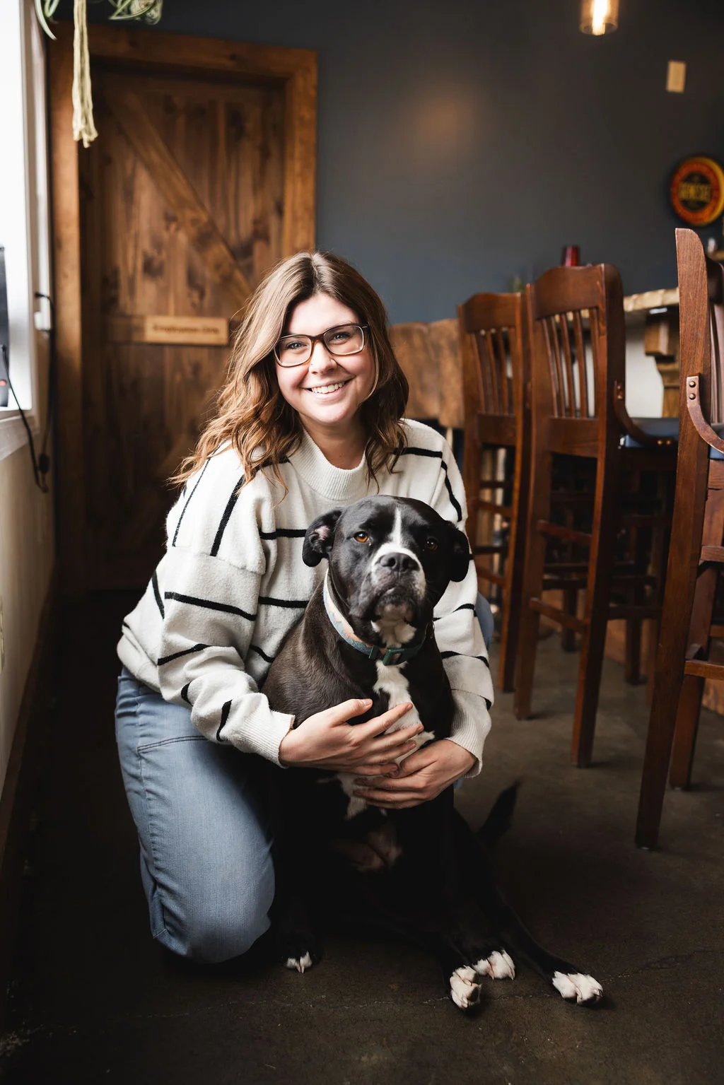 A person sitting on the floor next to a dog, smiling, in a cozy bar setting with wooden chairs in the background.
