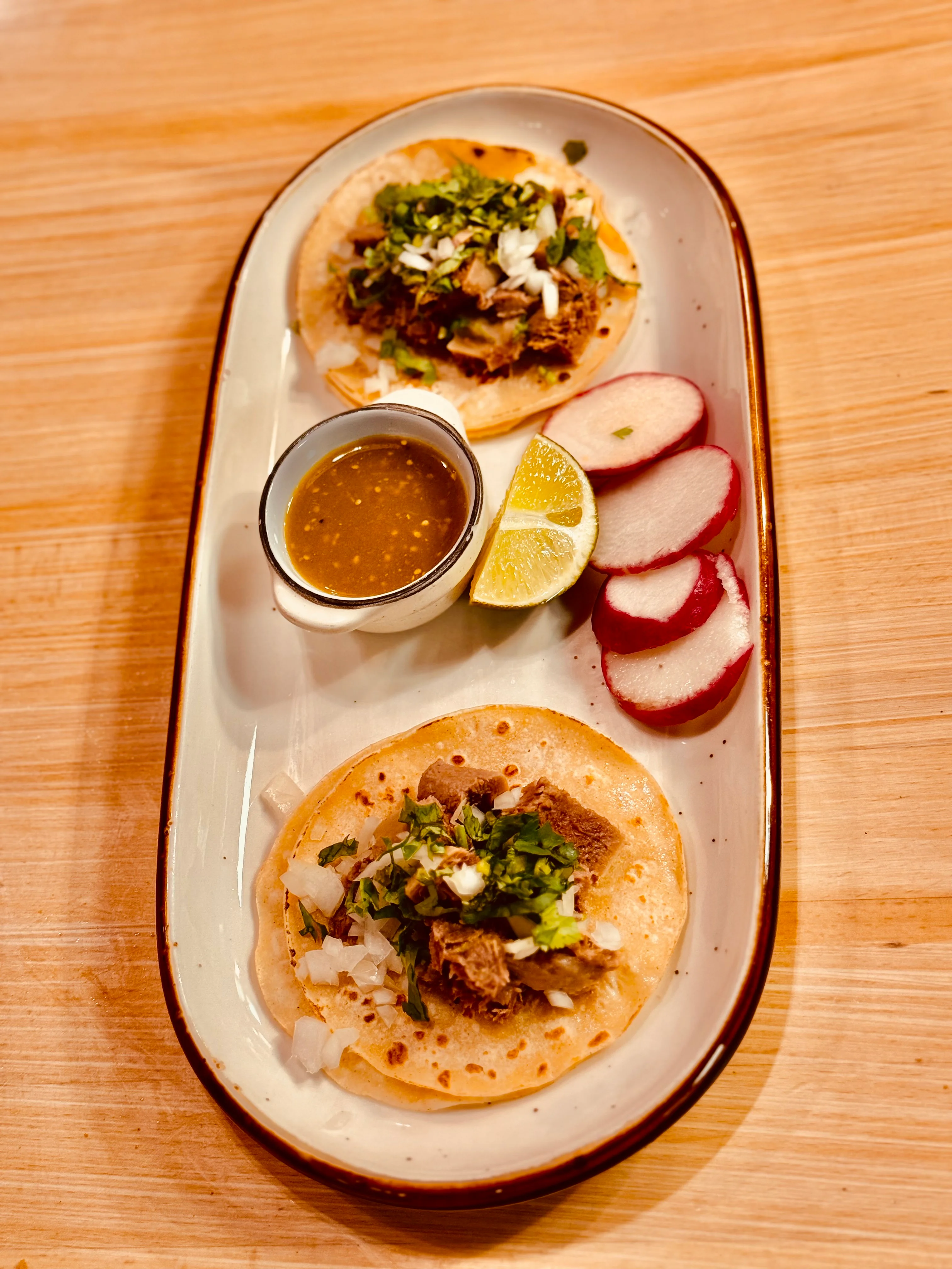 Two tacos filled with seasoned meat, garnished with onions and cilantro, served on a platter alongside lime wedges and radish slices with a bowl of sauce.