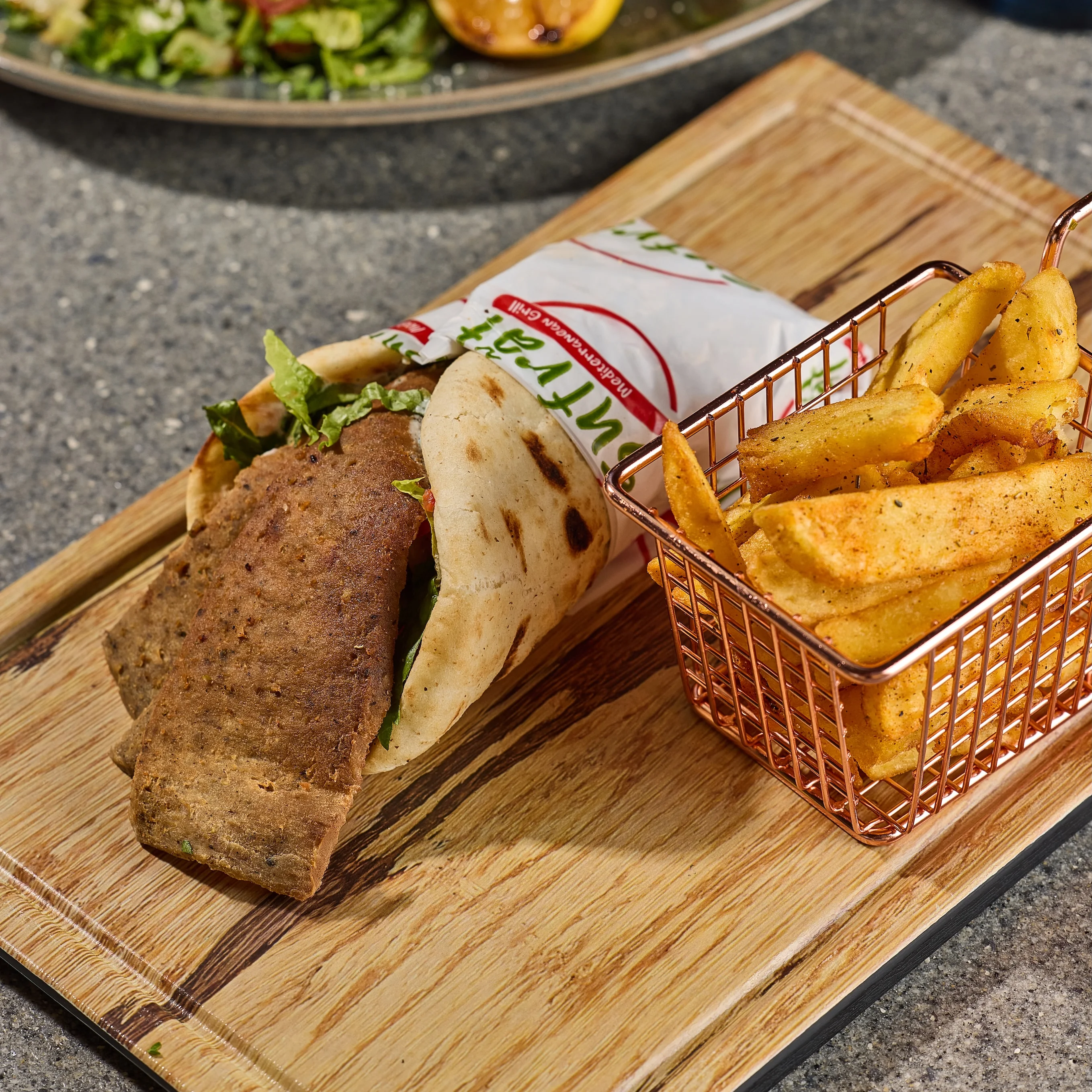 A gyro wrapped in flatbread with lettuce and roasted meat sits on a wooden board beside a small basket of crispy, golden fries.