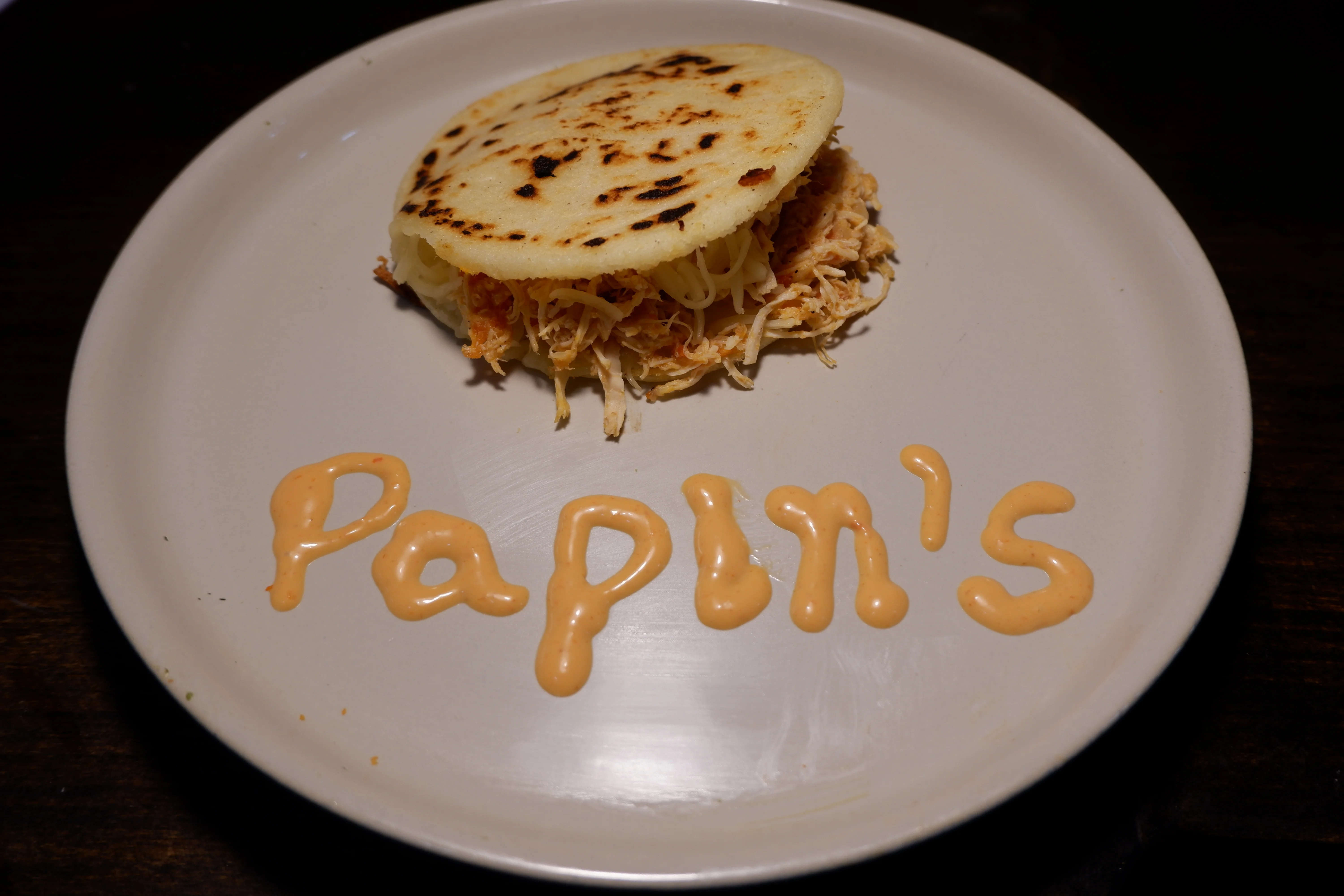 A handmade arepa filled with shredded chicken and cheese, placed on a plate with sauce spelling out a name.