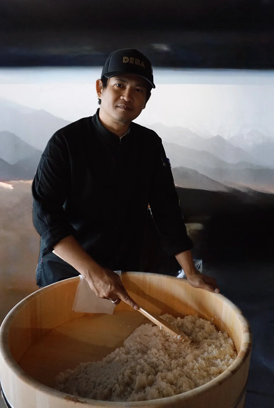Chef mixing rice in a wooden bowl with mountains in the background.