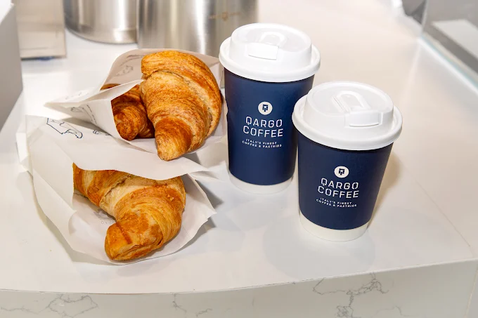 Two cups of coffee beside freshly baked croissants wrapped in paper, arranged on a light-colored surface.