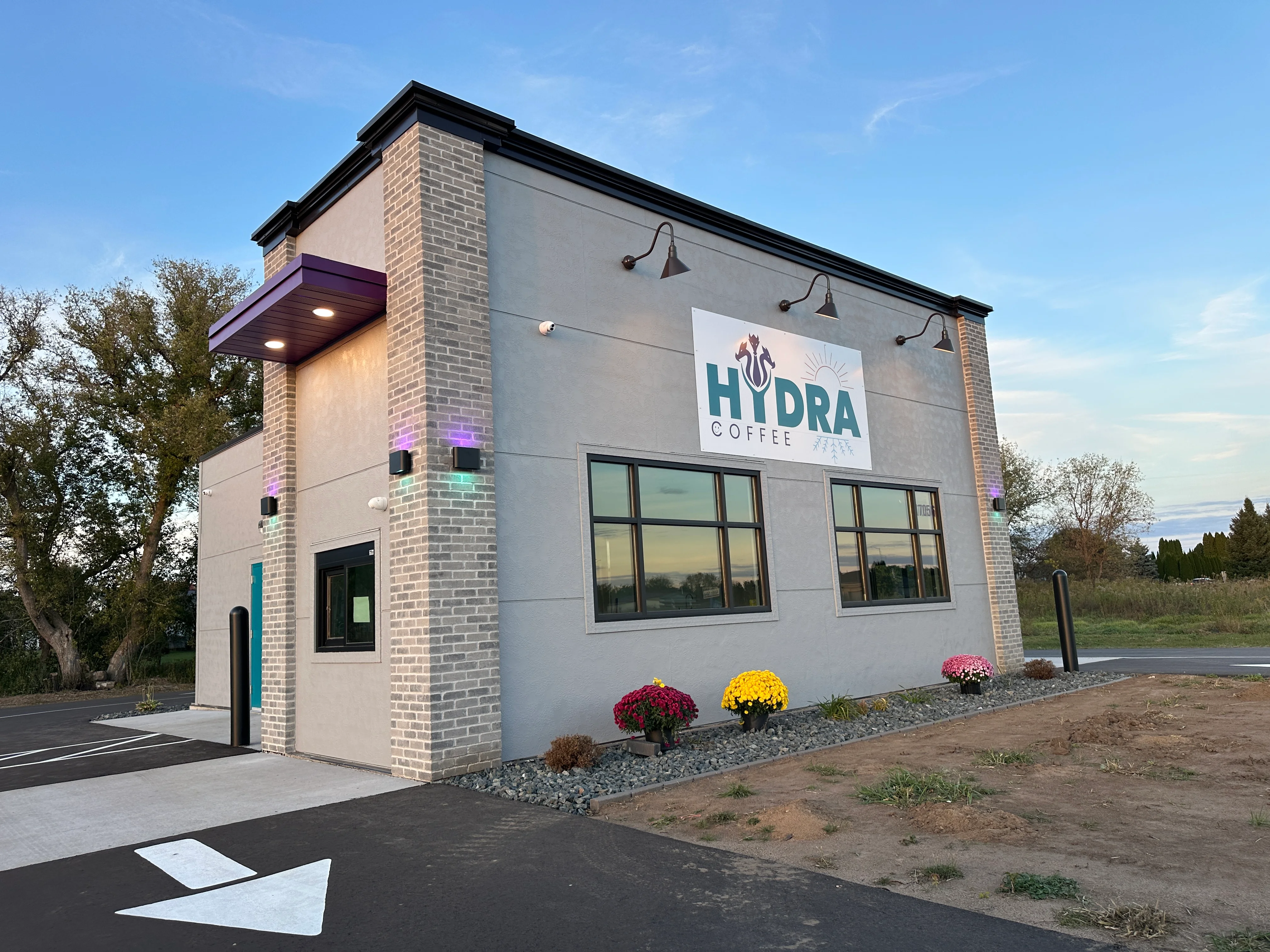 A modern coffee shop exterior with a sign, colorful plants, and a clear blue sky overhead.