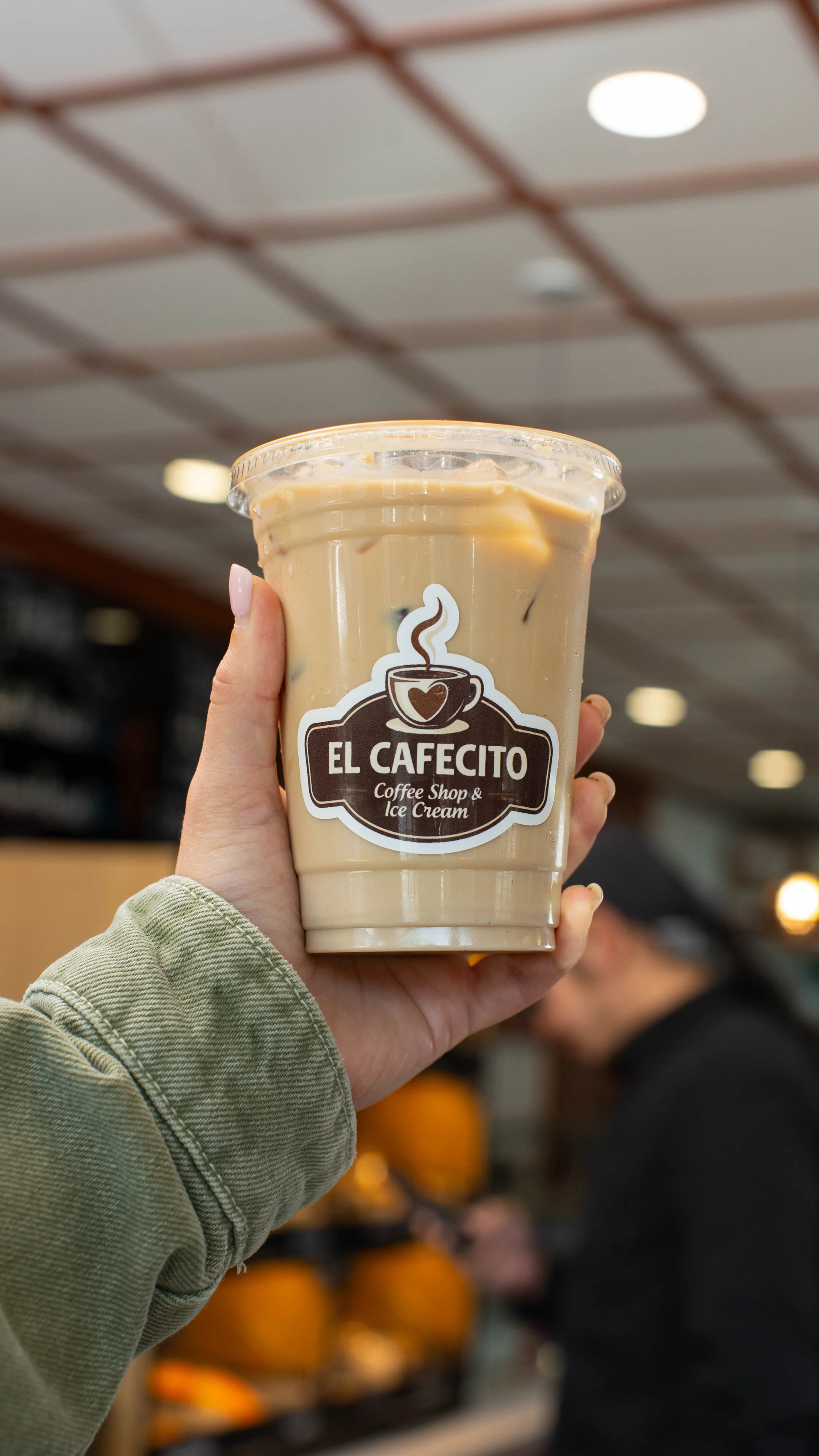 Iced coffee from El Cafecito held in a hand, with a cozy café background.