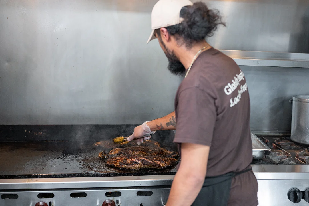 A chef wearing a baseball cap, a dark shirt, and an apron is seen in a professional kitchen, turning pieces of seasoned meat on a flat-top grill using tongs. Steam is rising from the cooking meat.