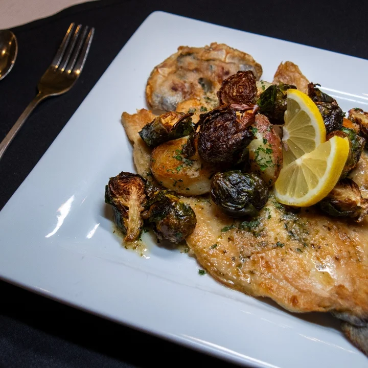 A grilled fish fillet served with roasted Brussels sprouts and baby potatoes, garnished with lemon slices and herbs.