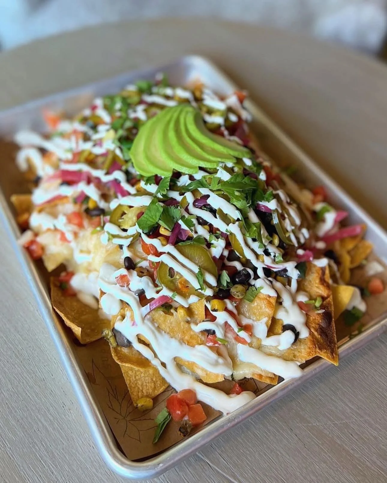 A platter of nachos topped with fresh ingredients including avocado slices, tomatoes, jalapeños, and drizzled with sour cream.