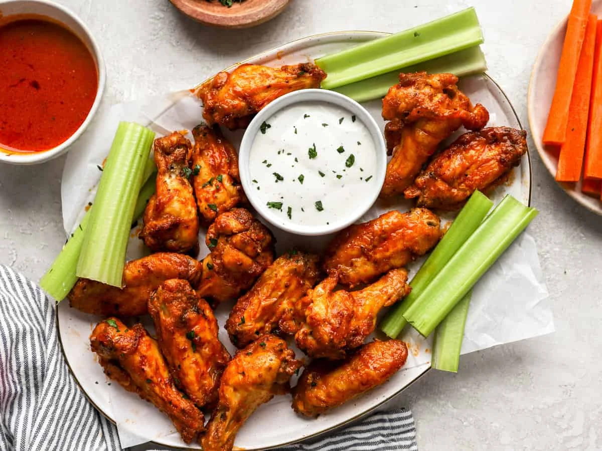 A platter of crispy chicken wings surrounded by celery sticks, served with a bowl of ranch dressing and hot sauce.