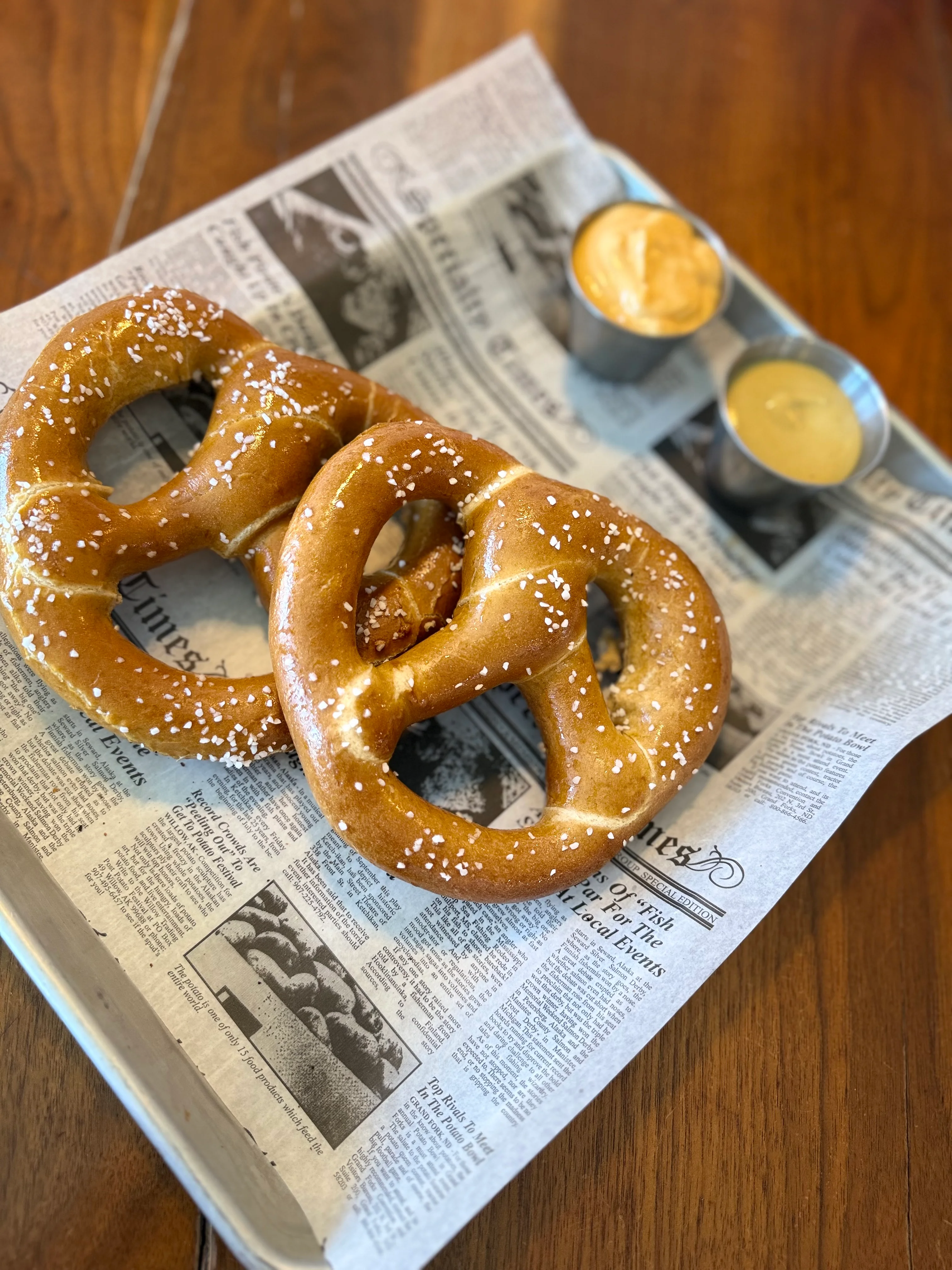 Two soft pretzels sprinkled with coarse salt, served with two dipping sauces of different colors on a newspaper-lined tray.