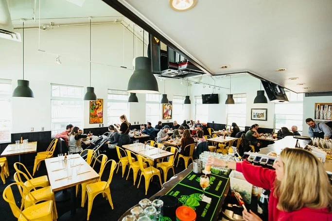 An interior view of a vibrant dining area featuring yellow chairs and wooden tables. Customers are seated and socializing, while a bartender prepares drinks at the counter.